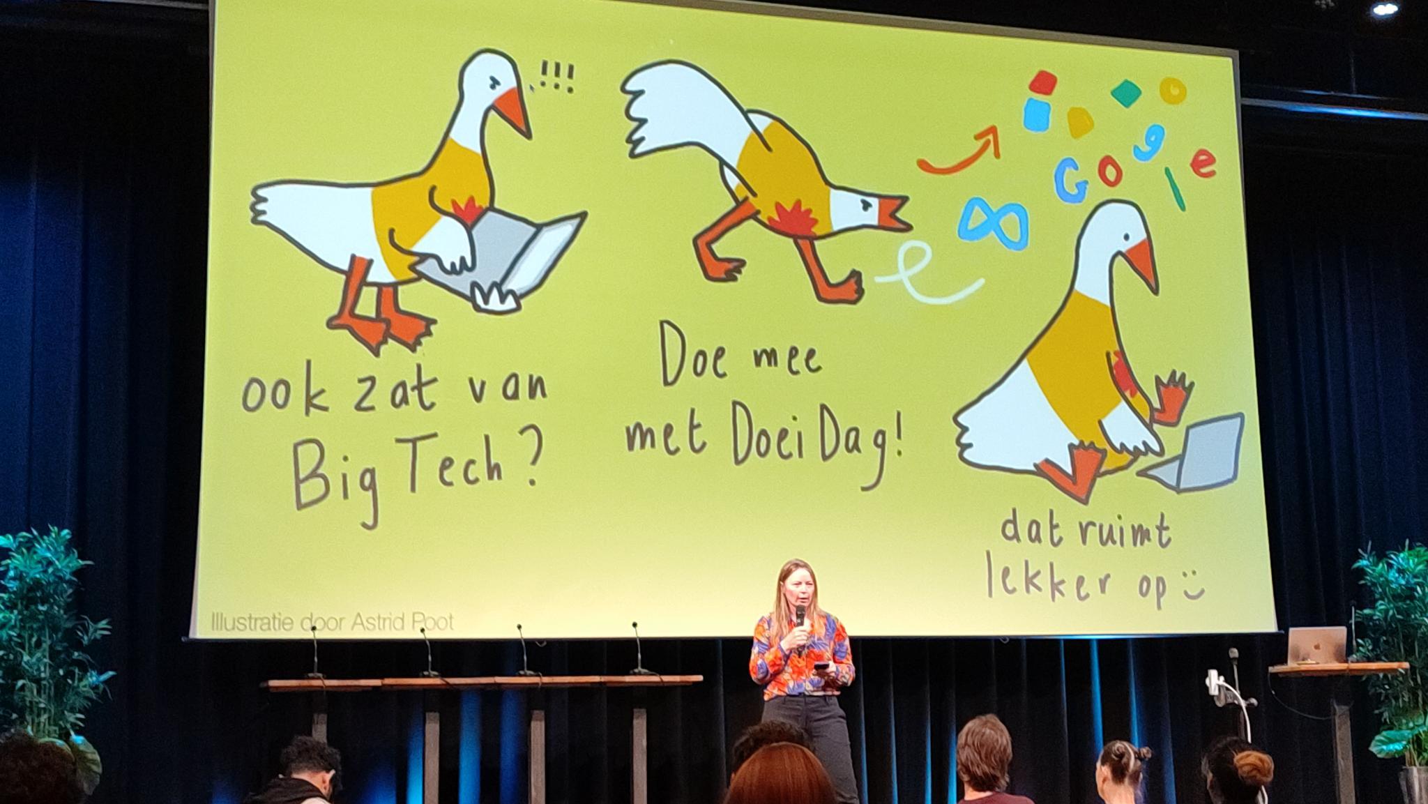 Foto van een enorm groot scherm met daarop de illustratie van Astrid Poot voor Doeidag. Met op de voorgrond, op een podium, Elmine die verteld over Doeidag tijdens de PublicSpaces conferentie 2026